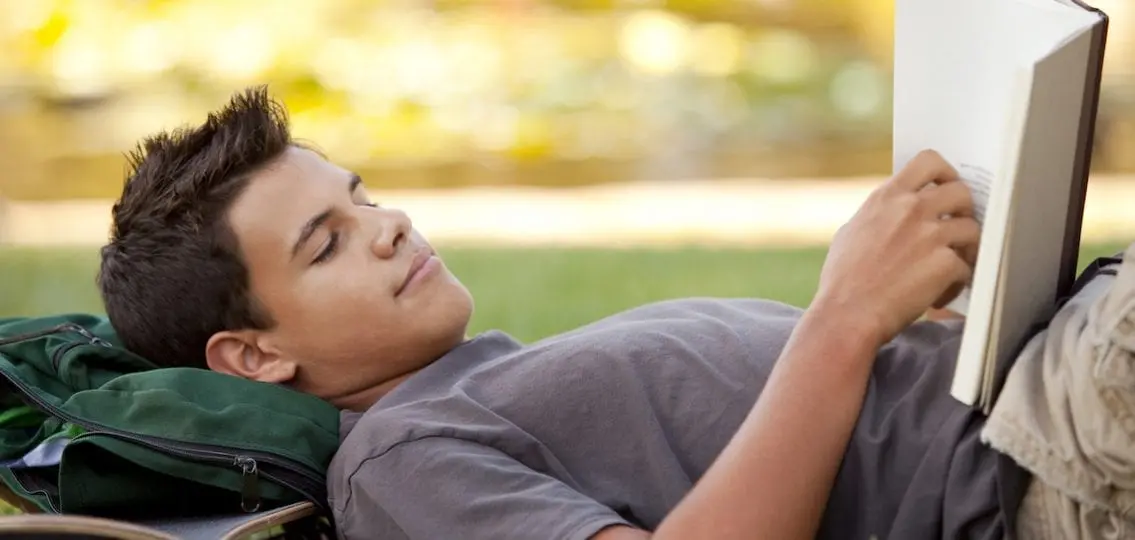 Teen boy reading a book while lying in the grass outside