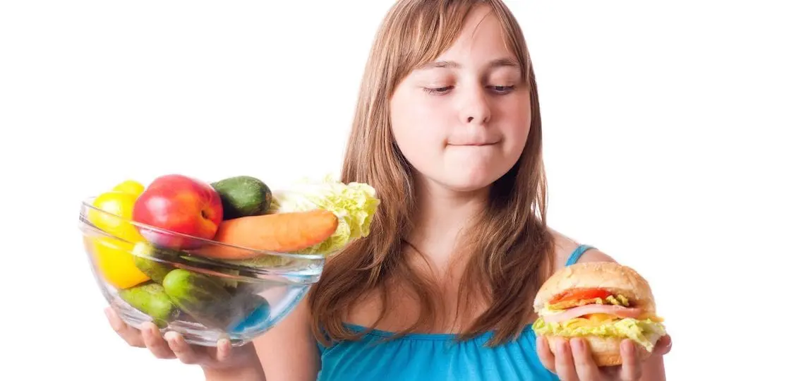 teenage girl debating between a bowl of healthy vegetables and fruits vs a cheeseburger while looking at the burger