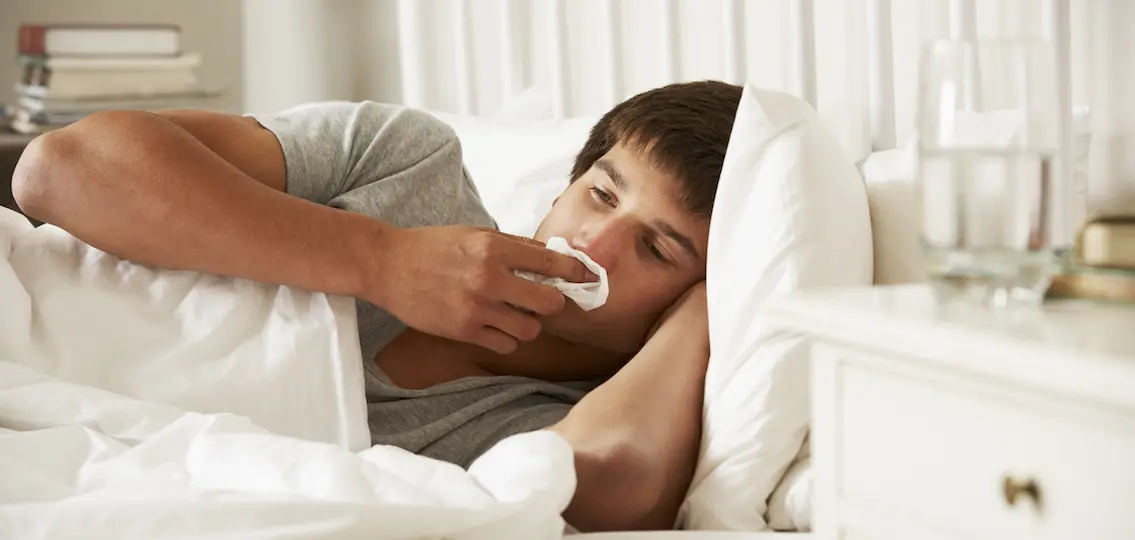 a sick teenage boy in his bed wiping his runny nose with a tissue