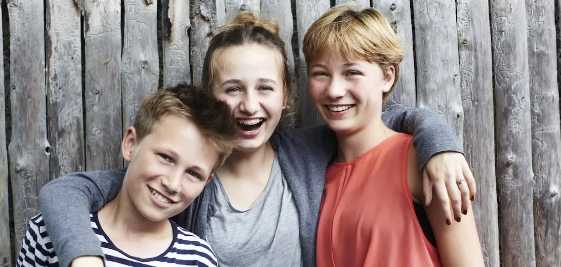 Three Siblings of varying ages smiling and hugging in front of a wooden fence