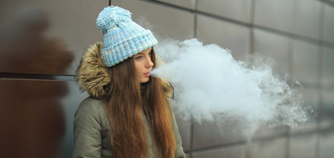 a teen girl breathing out either cigarette of vape smoke outside in the winter