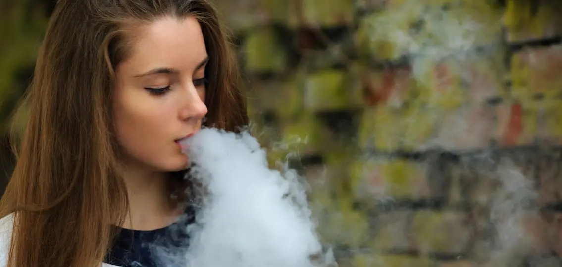 Teen girl blowing vape smoke with a brick wall behind her