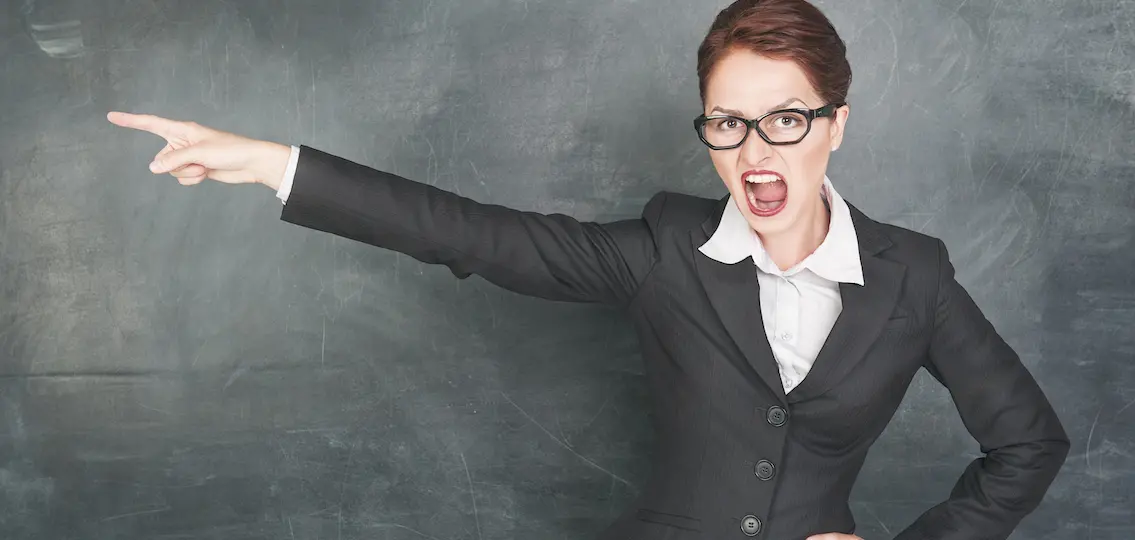 Angry teacher in glasses pointing out in front of blackboard