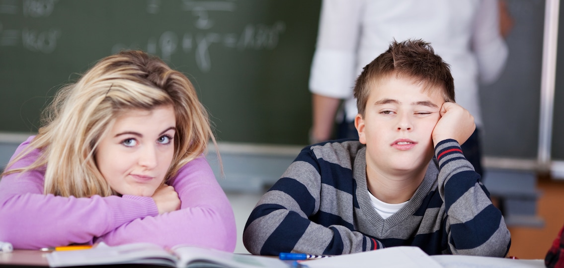 Bored students leaning on desk with teacher in background at classroom