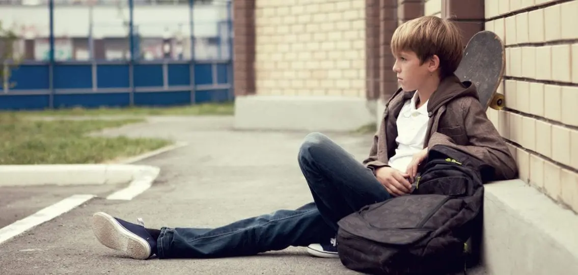 teen boy with a skateboard and a backpack leaning against a brick wall in a parking lot
