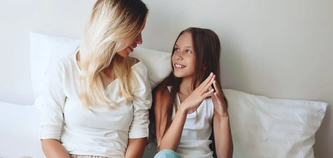 Mom and daughter sitting together on bed and smiling together