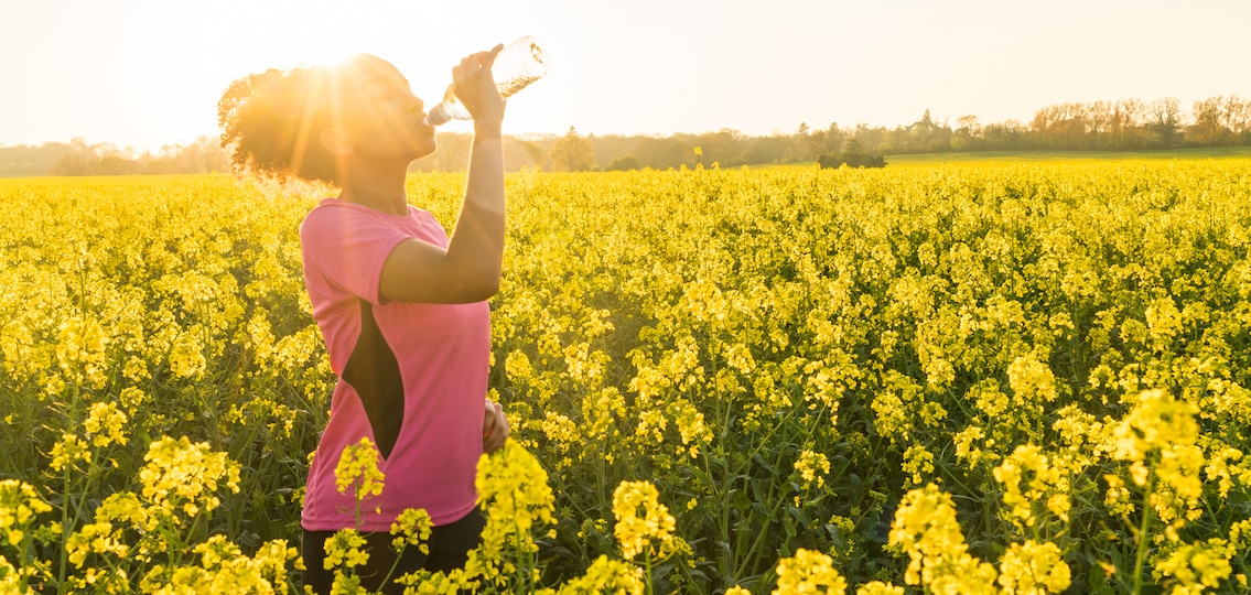 Outdoor portrait of beautiful teen girl athlete runner drinking water from a bottle in a field of yellow flowers at sunset in golden evening sunshine