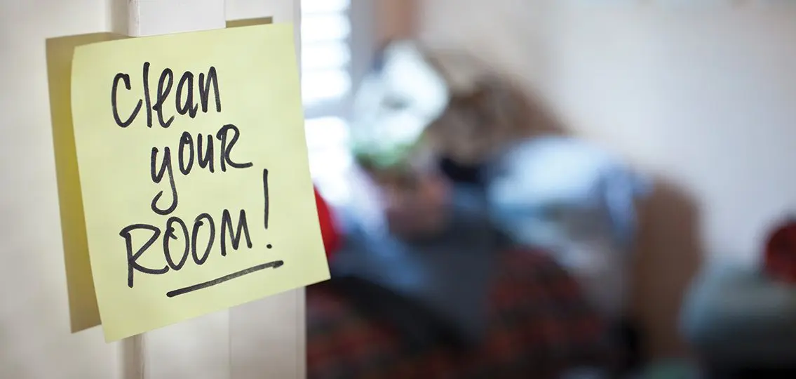 sticky note reading clean your room with blurry messy teen bedroom in background