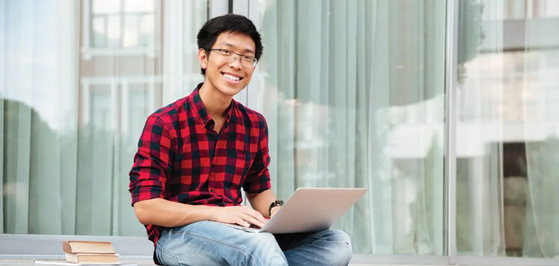 Happy asian young man in plaid shirt using laptop outdoors