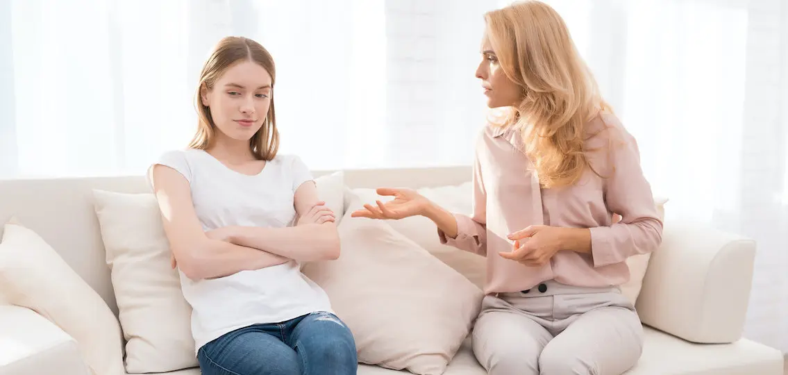 Mom and a teenage daughter are arguing with each other about daughter's behavior on a white couch in a white room