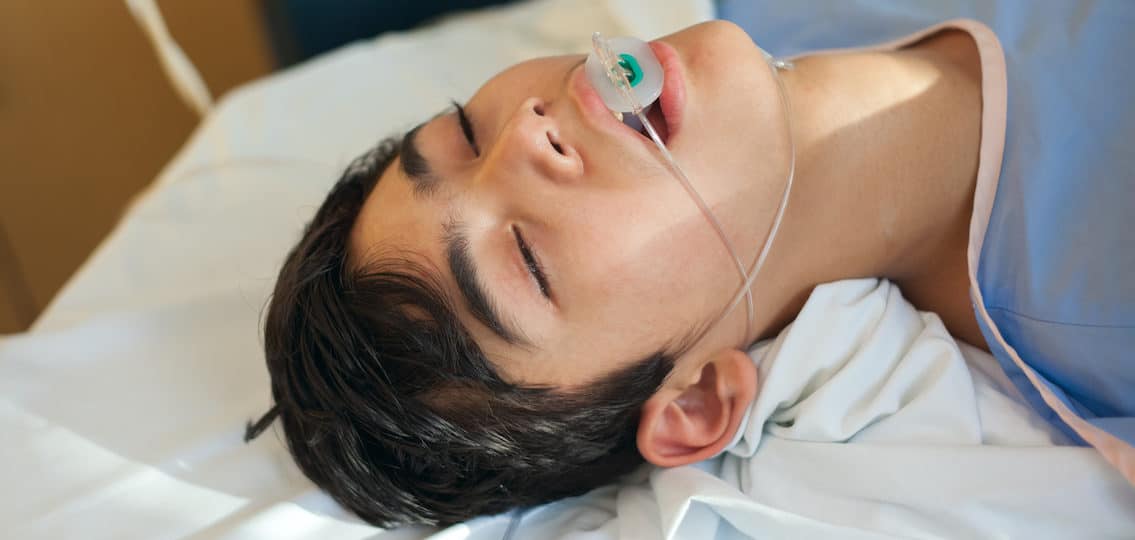 Young thirteen year old disabled biracial boy lying unconscious on hospital gurney bed in recovery room wearing blue hospital gown, breathing tube down throat after alcohol poisoning