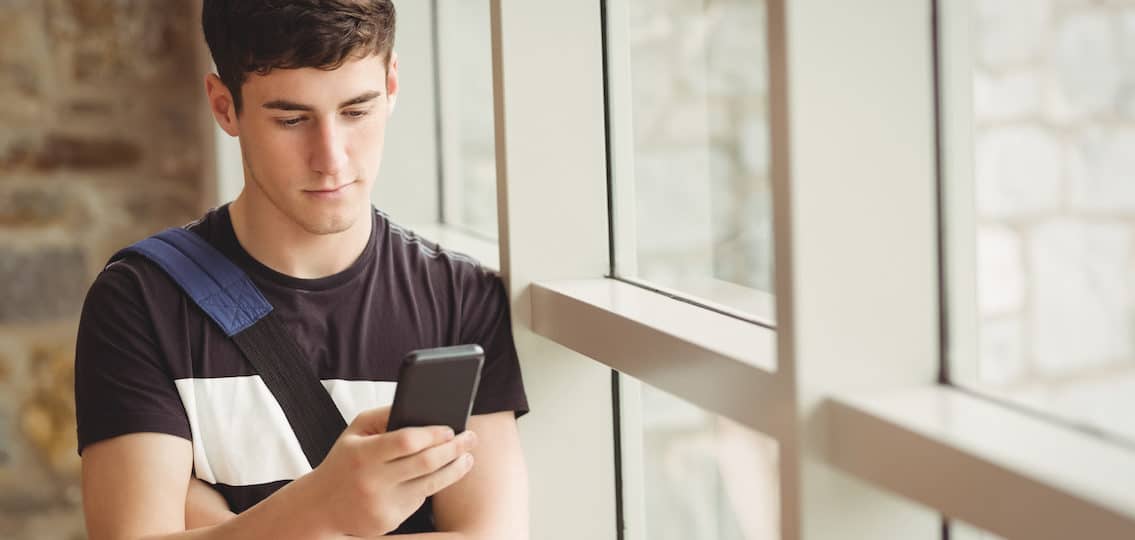 Male student waiting for text while leaning on window in college