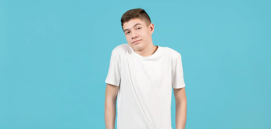 A teenager in a white T-shirt smiles uncertainly, shrugs. He stands on a blue background and his hands in his pockets.