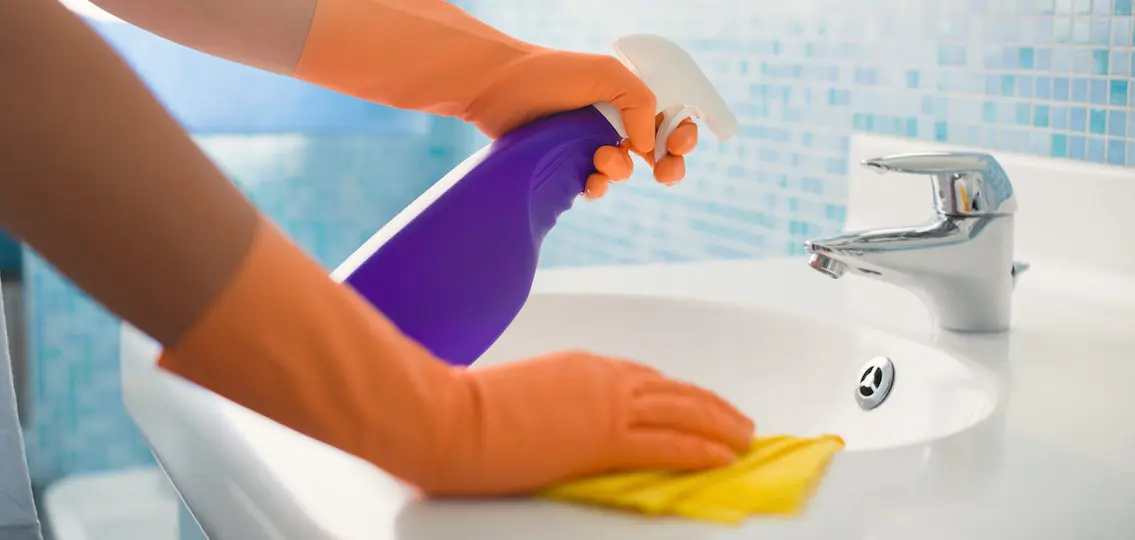 woman doing chores in bathroom at home, cleaning sink and faucet with spray detergent. Cropped view
