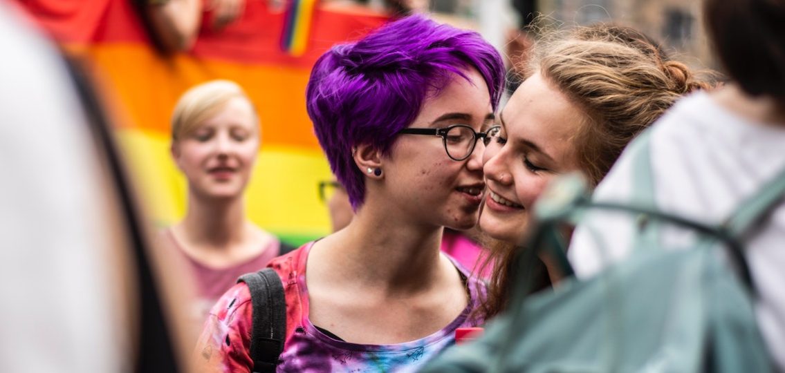 teen girl with purple hair kissing her girlfriend's cheek at a pride parade