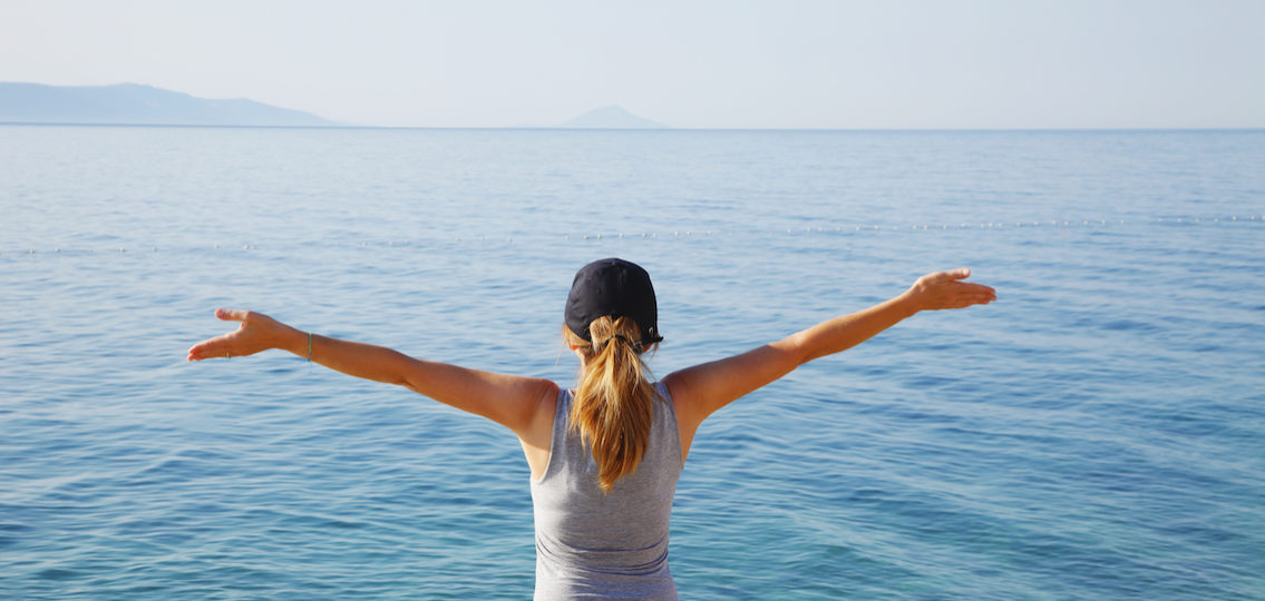 Rear view portrait of fitt mature woman standing with arms raised by the sea after workout while relaxing.