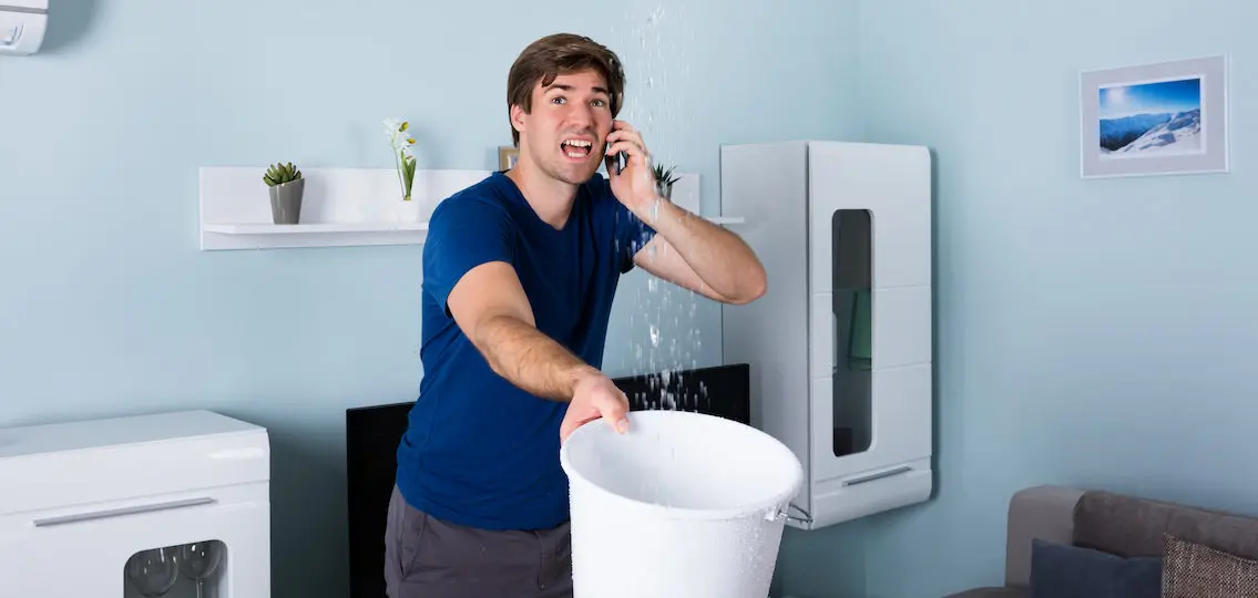 Worried college age man Calling Plumber While Leakage Water Falling Into Bucket At Home