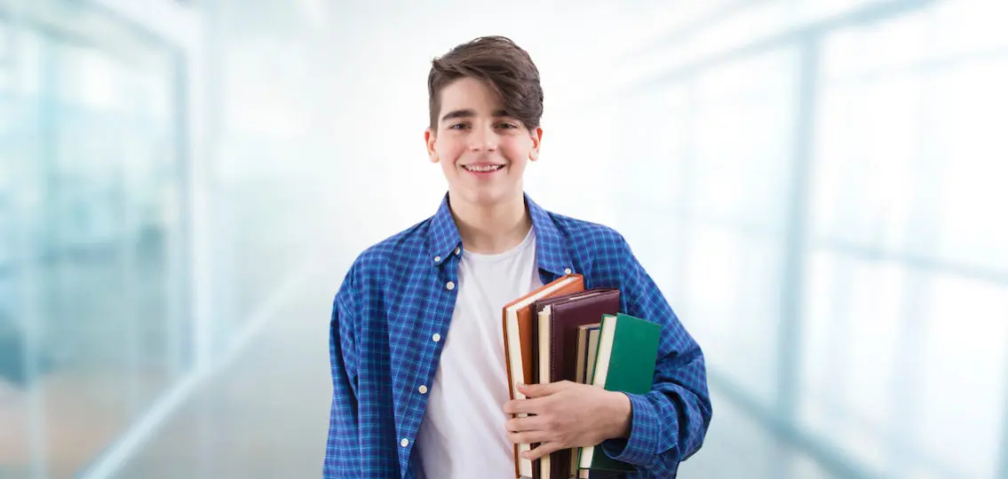 young teenage boy freshman holding books and smiling in school hallway