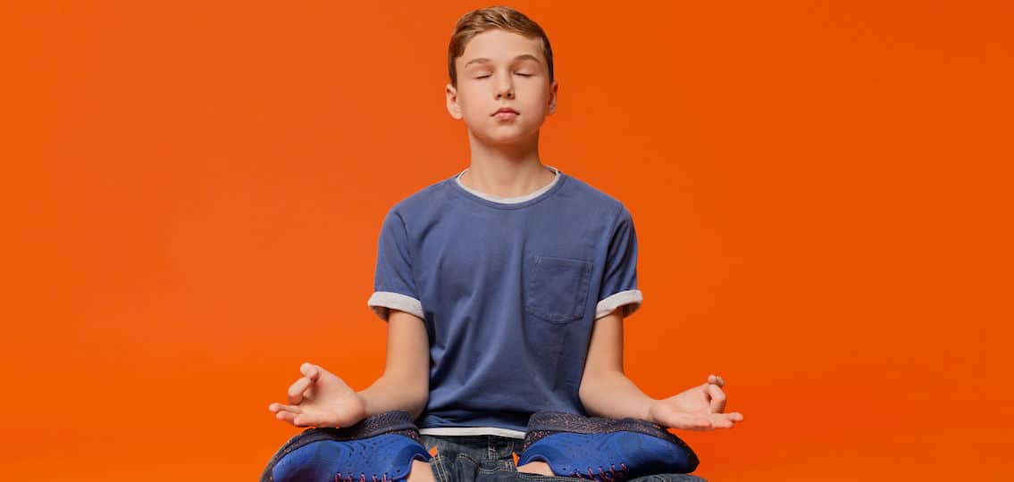 Calm boy sitting on floor and meditating in lotus position, practicing yoga, orange studio background
