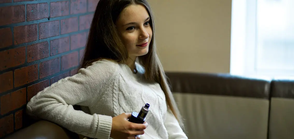 Vaping teenager. Young pretty teenage girl with problem skin smoking an electronic cigarette indoors. Life-threatening bad habit. Vape activity.