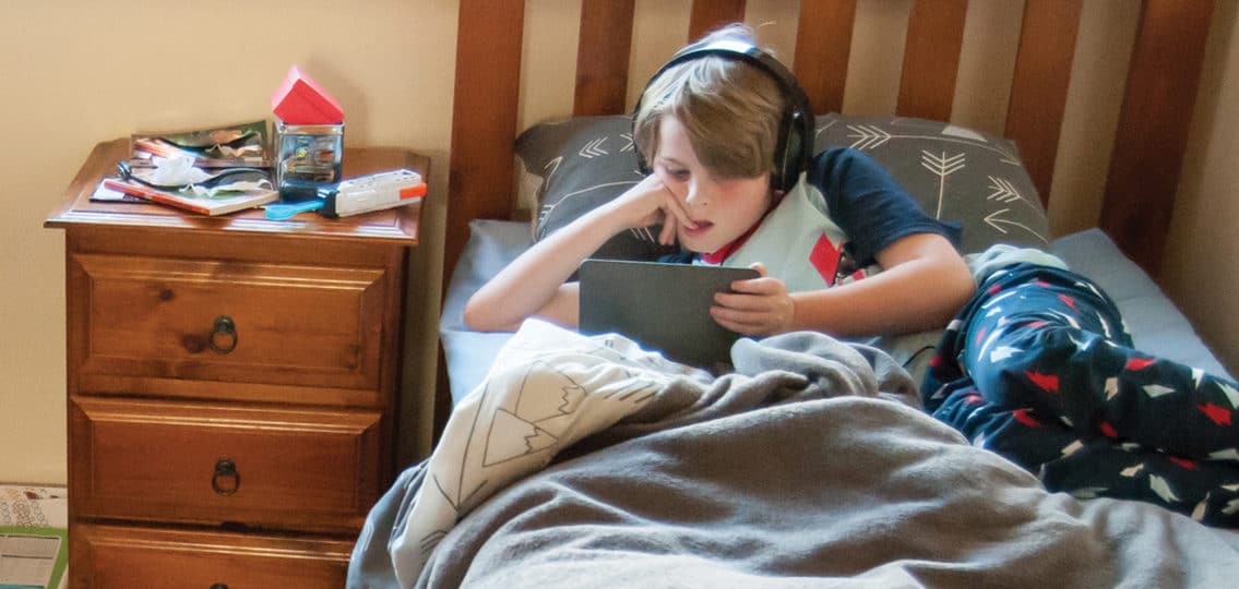 Child laying in bed on electronic device with a very messy room