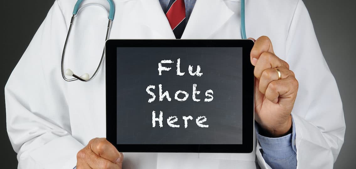 Closeup of a doctor holding a tablet computer with a chalkboard screen with the words Flu Shots Here. Man is unrecognizable.