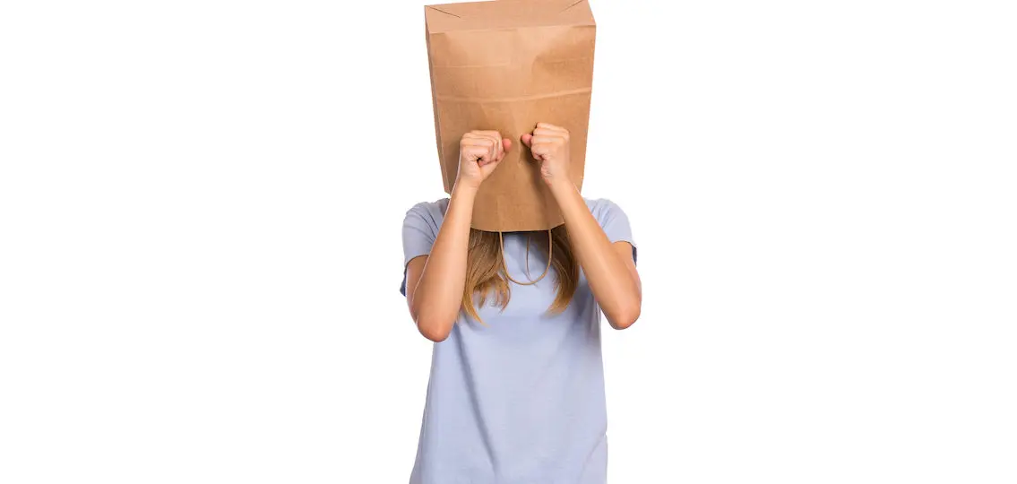 Unhappy teen girl with body dysmorphia wearing paper bag over head covering face with hands while crying. Upset teenager posing in studio. Child crying, not showing her tears.