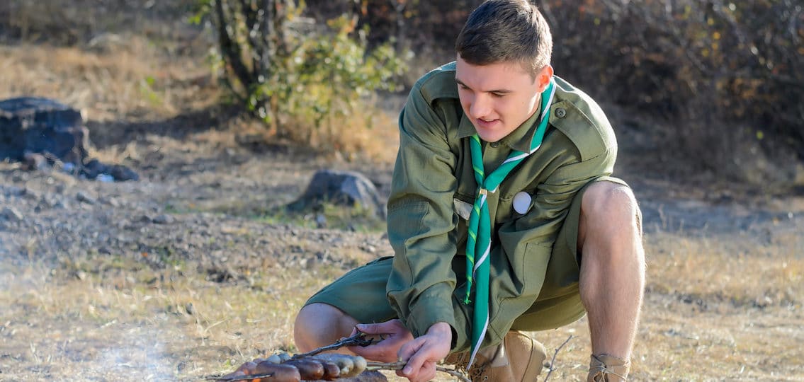 Teenage Eagle Scout in Uniform Crouching and Cooking Sausages on Sticks over Campfire