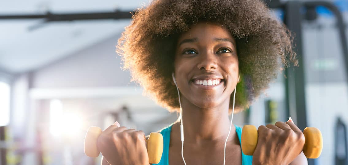 Happy healthy young African American teen girl working out in a gym with a pair of dumbbells and smiling during her weight training
