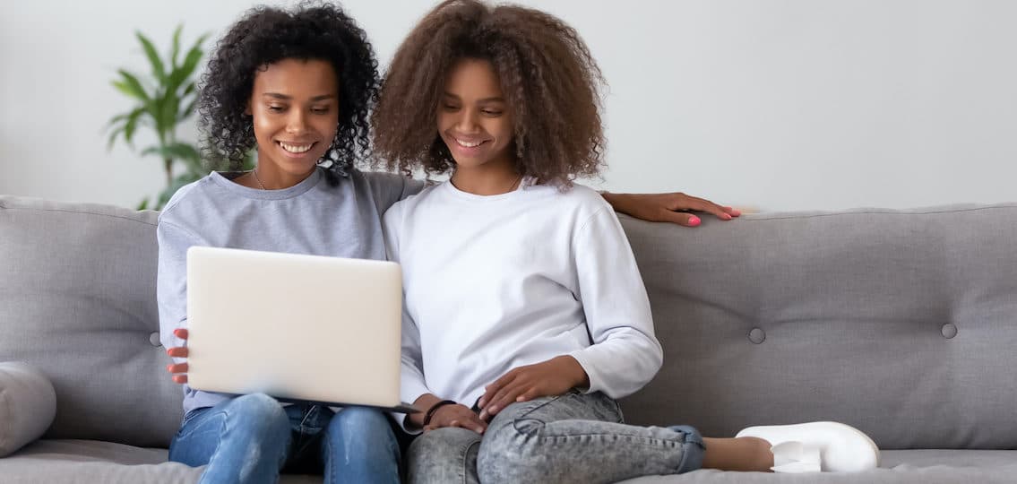 Happy african mother and teen daughter using laptop on sofa