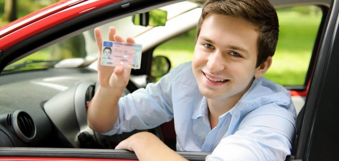 teen boy in drivers seat holding up learners permit and smiling
