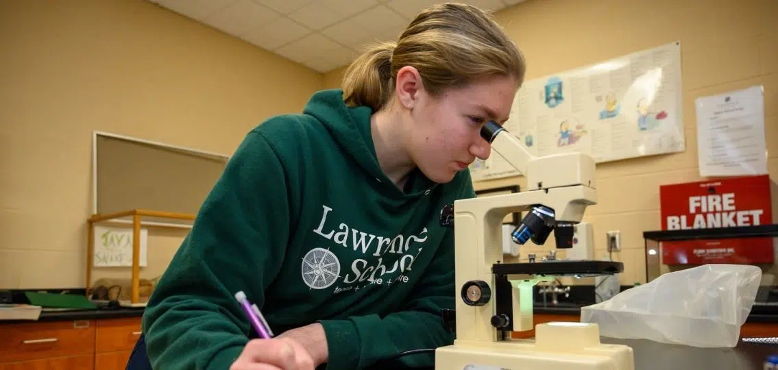 A teenager with a learning difference wearing a Lawrence School hoodie looking intro a microscope in a classroom and writing down her results