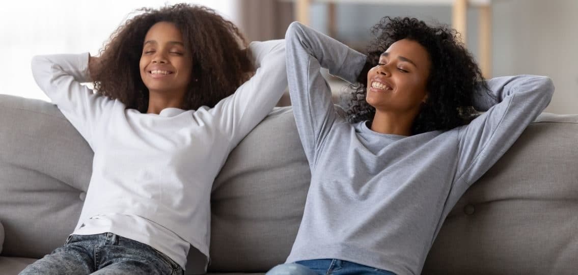 a teen girl and an adult woman matching clothes relaxing on a couch with their hands behind their heads smiling relaxing with a mental health day