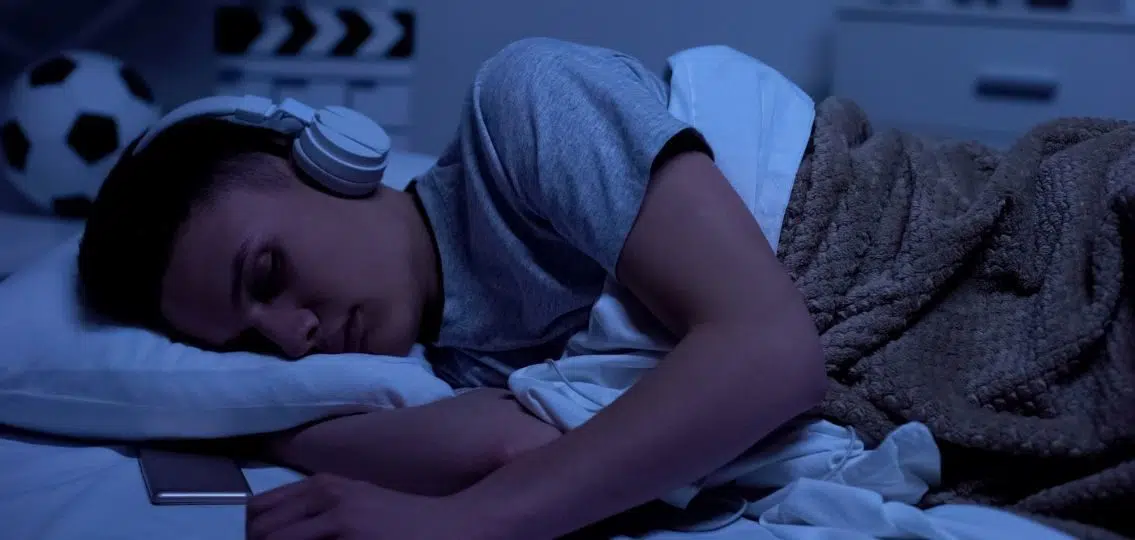 A teen boy asleep in his bed with his phone at night listening to music