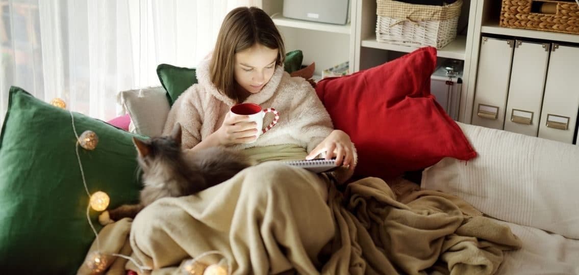 a teenager snuggled onto the couch on winter break with a hot chocolate a cat and a bunch of blankets