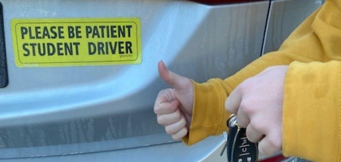 close up of teen giving a thumbs up with a pair of keys in front of a car with a please be patient student driver bumper sticker