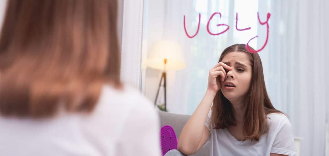teen girl suffering from poor body image because of negative body talk looking in a mirror that says ugly on it and crying