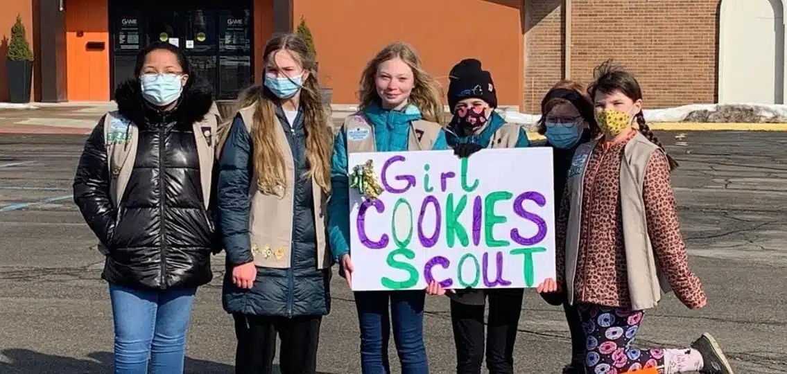 A group of Girl Scouts with a sign for girl scout cookies selling girl scout cookies outside