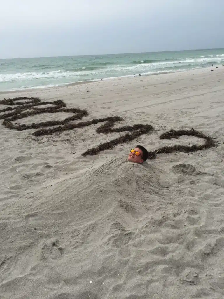 A funny picture of a boy buried in sand next to seaweed that spells out Prom?