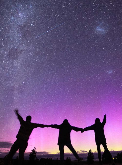 three people holding hands, silhouetted against a night sky filled with stars that they are facing