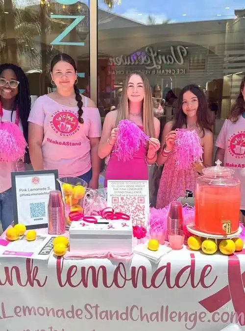 five teen girls dressed in pink standing behind a table with a pink lemonade breast cancer table cloth