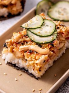 square piece of baked sushi made of rice and crab meat, topped with sliced cucumbers and spicy mayo