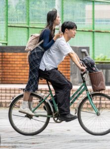 a boy riding a bicycle with a girl leaning on his back and standing on the hub of the back wheel