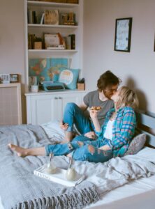 A man and woman sitting on a bed kissing. There is a tray holding drinks on the bed and the woman is holding a slice of pizza.